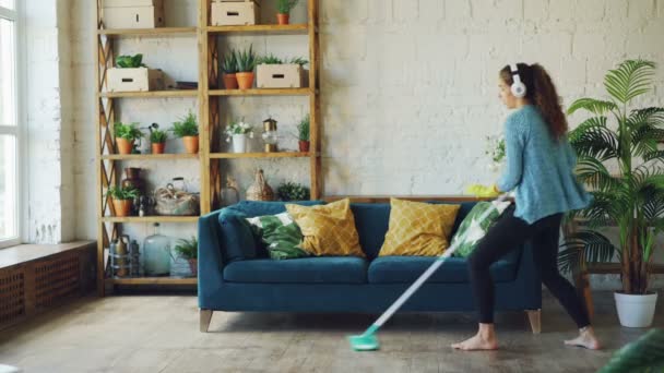 Glad ung kvinna dansar med mopp vid rutinmässiga sanering i trevligt loft stil lägenhet, hon lyssnar på musik med trådlösa hörlurar och sång. — Stockvideo