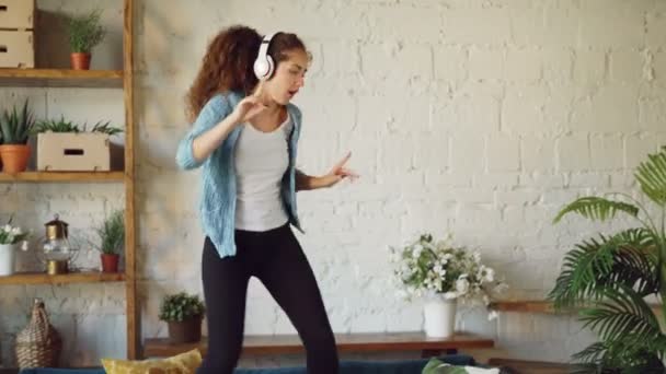 Morena atraente está se divertindo em casa ouvindo música através de fones de ouvido, cantando e dançando pulando no sofá. Conceito de tecnologia moderna e de jovens . — Vídeo de Stock