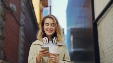 Mutlu kız ceket ve holding take-away kahve sokakta yürürken smartphone kullanarak ağır çekim. Kentsel yaşam tarzı, millennials ve genç insanlar kavramı.
