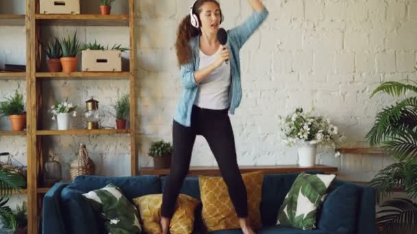 Happy young woman is listening to music in headphones, dancing on sofa and singing in hairbrush like in microphone, girl is having fun in free time enjoying song. — Stock Video