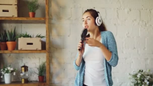 Грайлива дівчина співає в щітці для волосся і танцює на дивані, слухаючи музику в бездротових навушниках. Час дозвілля весело, щасливі тисячоліття і концепція технології . — стокове відео