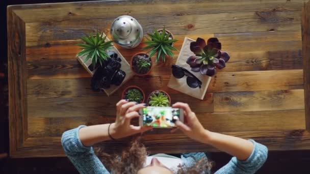 創造的な写真家が木製のテーブルの上の植物、カメラ、サングラスのフラット横たわっていた写真を撮ることのスマート フォンを使って、女性が画面に触れると撮影. — ストック動画