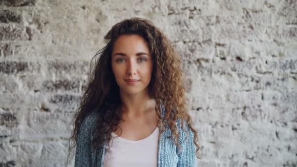 Portrait de belle jeune femme avec de longs cheveux bouclés debout contre un mur de briques, souriant et regardant la caméra. Concept attrayant pour les personnes, les intérieurs et les jeunes . — Video