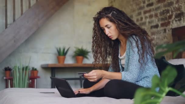 Νεαρή γυναίκα πληρωμή κρατώντας τραπεζική κάρτα και να εργάζονται με το laptop κάθεται στο κρεβάτι στο σπίτι. Αγοράζουν πράγματα σε απευθείας σύνδεση, internet και σύγχρονη τεχνολογία έννοια. — Αρχείο Βίντεο