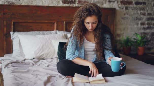 Menina bonita ler livro sentado na cama em casa e segurando copo com bebida desfrutando de tempo livre e descanso. Jovens, hobby e conceito de interior moderno . — Vídeo de Stock