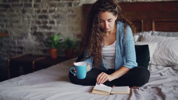 Aantrekkelijke jonge vrouw met donker krullend haar is lezen fotoboekpagina draaien en genieten van literatuur zittend op bed met kopje thee. Ontspannen mensen, hobby en huizen concept. — Stockvideo