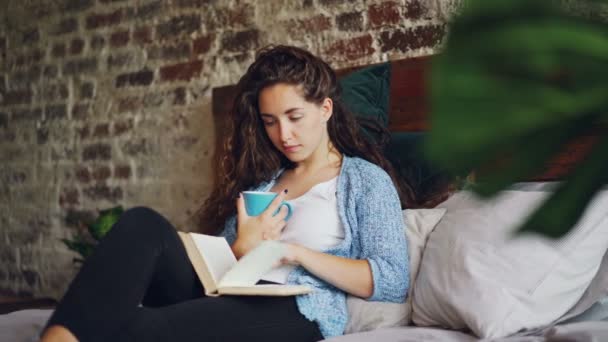 Ragazza allegra sta leggendo il libro divertente in camera da letto sdraiato sul letto comodo, sorridente e in possesso di tazza con bevanda. Hobby, persone felici, splendidi interni e concetto di letteratura . — Video Stock