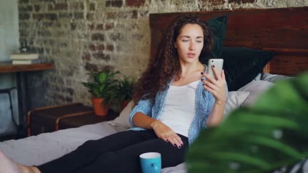 Menina feliz está conversando on-line usando smartphone olhando para a câmera do telefone, falando em sorrir sentado na cama no quarto no apartamento moderno. Conceito de tecnologia e conversação . — Vídeo de Stock