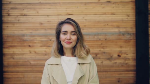 슬로우 모션 카메라를 보고 다음 야외 코트를 입고 서 서 웃 고 웃 고 금발 머리를 가진 행복 한 젊은 여자의 초상화. 사람과 감정 개념. — 비디오