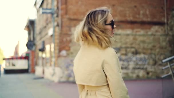 Retrato en cámara lenta de una atractiva chica rubia caminando por la calle, sonriendo y mirando a la cámara. Gente alegre, ropa hermosa y concepto de estilo de vida urbano . — Vídeo de stock