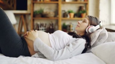Çekici hamile kadın evde yatakta yatarken ve karnı okşayarak kulaklık ile müzik dinliyor. Eğlence, mutlu hamilelik ve ev kavramı.