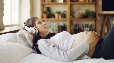 Bekliyor Anne karnı okşayarak ve kulaklık ev yatak odasında yatakta yalan söylemekte rahatlatıcı ile müzik dinlemek. Modern teknoloji, gevşeme ve gebelik kavramı.