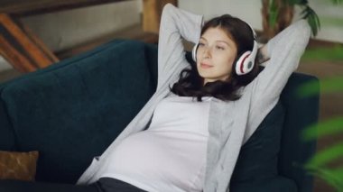 Güzel kız anne kulaklıklar giyen ve evde kanepede rahatlatıcı müzik dinlemek bekliyor dinlenme ve şarkıları zevk. Aile, gevşeme ve mutlu hamilelik kavramı.