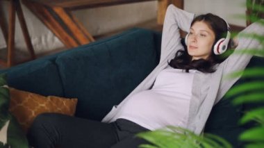 Hamile genç kadının evde oturma odasında kanepede oturan kablosuz kulaklık ile müzik dinliyor. Güzel bir kadın, modern teknoloji ve iç kavramı.