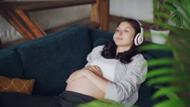 Hamile Giyim çekici hamile kadın kanepede yatan, karnı okşayarak ve kulaklık ile müzik dinlemek. Gevşeme, Aile ve modern yaşam kavramı.