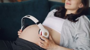 Gülümseyen hamile kadın bebeği için aşk ifade karnı büyük kulaklıklar ile rahat koltukta yatıyor. Modern teknoloji ve annelik kavramı.