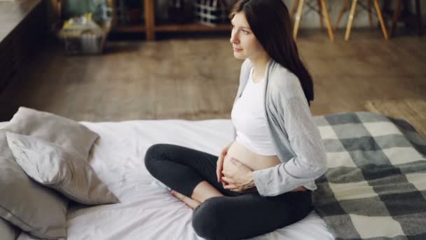 Schwangere streichelt ihren Bauch, sitzt zu Hause auf einem Doppelbett und genießt Ruhe und Freizeit. modernes Interieur, werdende Mütter und Konzept der Zärtlichkeit. — Stockvideo