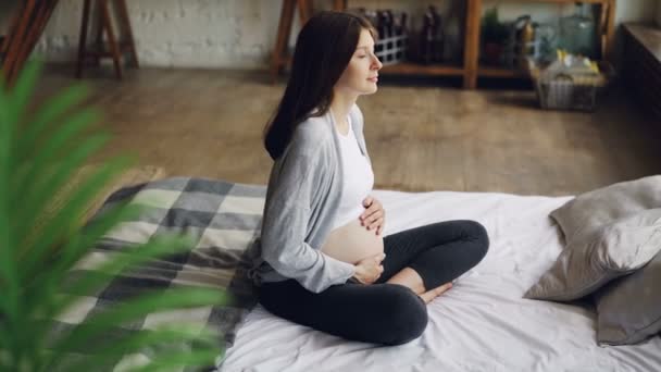 Schattig verwacht moeder sluit haar ogen en streelde haar buik gevoel baby zittend op bed in modern huis. Zwangere vrouwen, tederheid en liefde concept. — Stockvideo