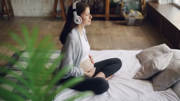 Hermosa mujer embarazada está acariciando su vientre sentado en la cama en un apartamento moderno y escuchando música con auriculares. Embarazo, interiores y concepto de felicidad . — Vídeo de stock