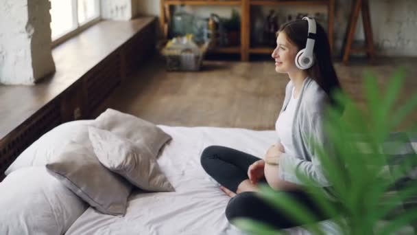 Улыбающаяся беременная девушка слушает музыку в наушниках, ласкает живот и расслабляется, сидя на двуспальной кровати в современной квартире. Концепция людей, семьи и домов . — стоковое видео