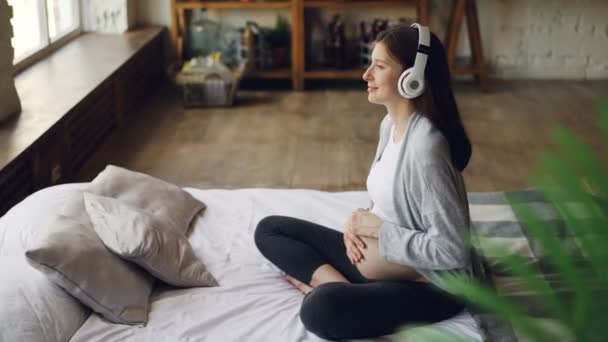 Feliz esperando mãe está acariciando sua barriga e ouvir música através de fones de ouvido descansando na cama de casal em casa moderna agradável. Conceito de família, interiores e tecnologia . — Vídeo de Stock