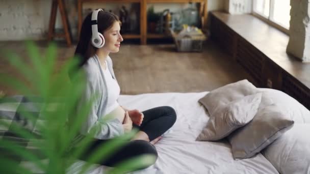 Alegre joven embarazada está acariciando su vientre y escuchando música disfrutando de una hermosa canción con auriculares y mirando por la ventana sentada en la cama en casa . — Vídeo de stock