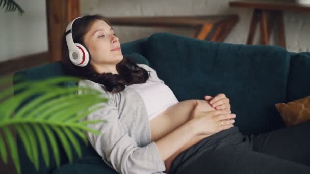 Verwacht moeder is ontspannend op Bank, luisteren naar muziek met een koptelefoon en streelde haar buik met liefde en zorg. Aantrekkelijke vrouwen, familie en vrije tijd concept. — Stockvideo