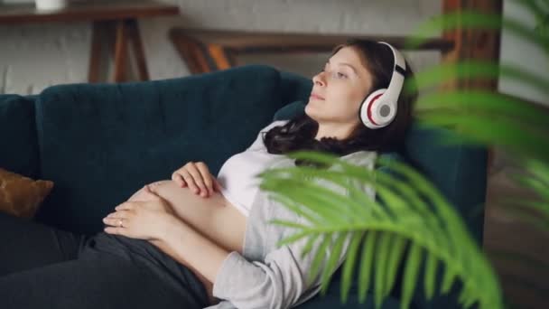 Jovem grávida com cabelo escuro está ouvindo música usando fones de ouvido sem fio modernos e acariciando sua barriga expressando amor. Conceito de maternidade, tecnologia e casa . — Vídeo de Stock