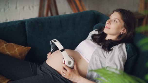 Kochający, spodziewając się matka trzyma bezprzewodowe słuchawki na jej brzuch, leżąc na kanapie w domu i odpoczynku. Nowoczesna technologia, kochających rodziców i koncepcja muzyki. — Wideo stockowe
