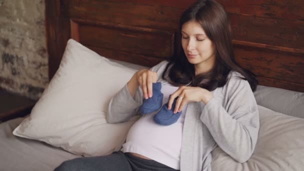 Šťastná mladá žena si hraje s baby boty chůze na její těhotné břicho a usmívá se přemýšlet o dítěti s láskou a něhou. Těhotenství a štěstí koncepce. — Stock video