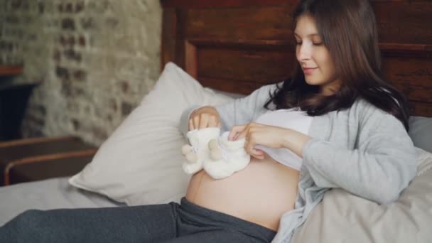 Embarazada jovencita está sosteniendo adorables zapatos de bebé en las manos y dando pequeños pasos sobre su vientre jugando con el feto mirando su barriga con amor . — Vídeo de stock