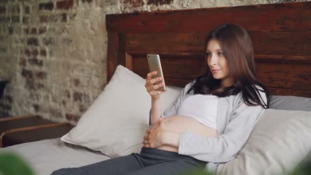 Jolie jeune femme qui attend sa mère utilise un smartphone, caressant son ventre et souriant couché dans son lit à la maison. Technologie moderne, grossesse et concept humain . — Video