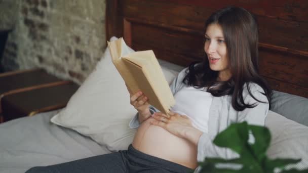 La joven embarazada está leyendo un libro en voz alta y acariciando su protuberancia con amor y ternura, la chica sonriente está disfrutando de la literatura y el descanso. Concepto de embarazo y hobby . — Vídeos de Stock