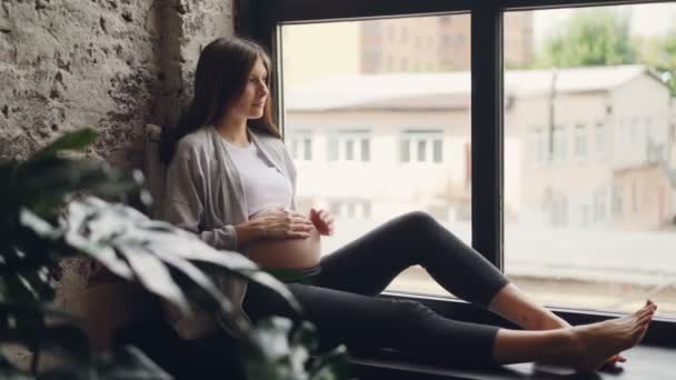 Chica embarazada hermosa morena está sentada en el alféizar de la ventana, acariciando su vientre y mirando por la ventana descansando. Interiores modernos, embarazo y concepto de personas . — Vídeos de Stock