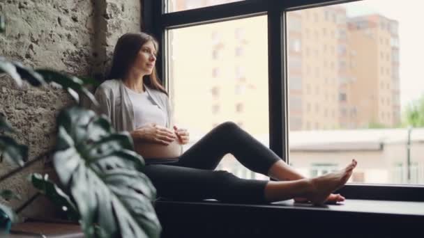 Atractiva mujer embarazada está sentada en el alféizar de la ventana y acariciando su bulto del bebé, ella está mirando hacia fuera y luego cerrando los ojos y descansando. Interiores modernos y concepto de embarazo . — Vídeo de stock