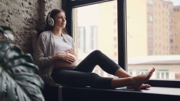 Oldukça hamile kadının karnı okşayarak ve pencere pervazına ev giyiyor rahat hamile giyim, oturan kulaklıklar üzerinden müzik. — Stok video