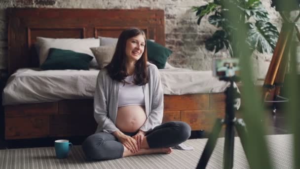 Beautiful young woman blogger is recording video about pregnancy for her vlog sitting at home on the floor, talking and looking at smartphone camera on tripod. — Stock Video