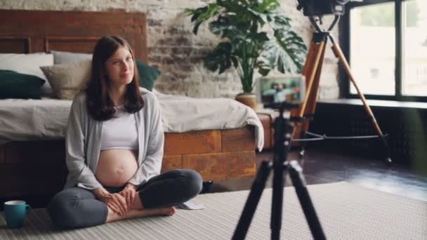 Donna incinta blogger femminile sta registrando video per i seguaci sulla gravidanza, ragazza è seduta sul pavimento della camera da letto, parlando e sorridendo utilizzando smartphone su treppiede . — Video Stock