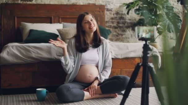 Bella giovane signora in attesa di madre e popolare vlogger sta registrando video per blog online seduto sul pavimento in appartamento moderno e parlando guardando la fotocamera dello smartphone . — Video Stock