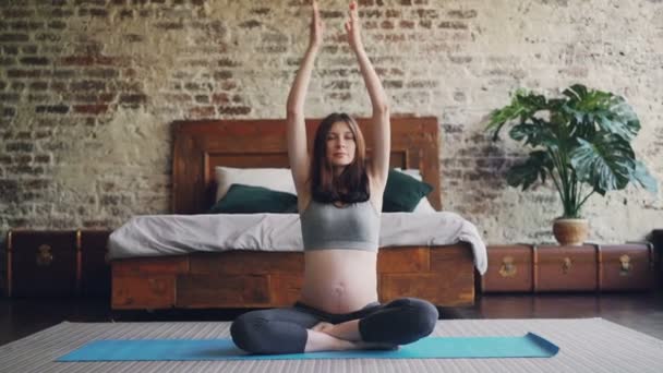 Dolly toma de bonita madre embarazada practicando yoga en casa sentada en la estera en el piso del dormitorio en pose de loto, levantando los brazos y luego poniendo las manos sobre las rodillas . — Vídeo de stock