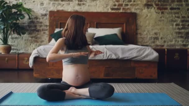 Adatta ragazza incinta sta facendo esercizi sportivi seduti sul tappeto yoga sul pavimento della camera da letto e girando il suo corpo a destra ea sinistra. Stile di vita sano, interni moderni e concetto di gravidanza . — Video Stock