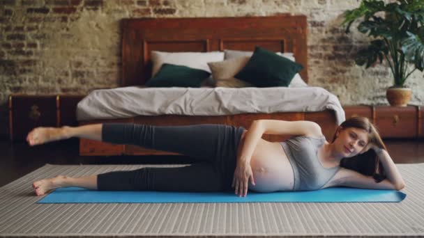 La mujer embarazada en forma está levantando su pierna acostada en la esterilla de yoga en el piso del dormitorio haciendo ejercicios deportivos. Estilo de vida saludable, esperando el concepto de madres e interiores . — Vídeos de Stock