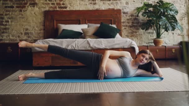 Attractive young lady expecting mother is doing sports lying on mat and raising leg up practising alone at home in bedroom. People, health and well-being concept. — Stock Video