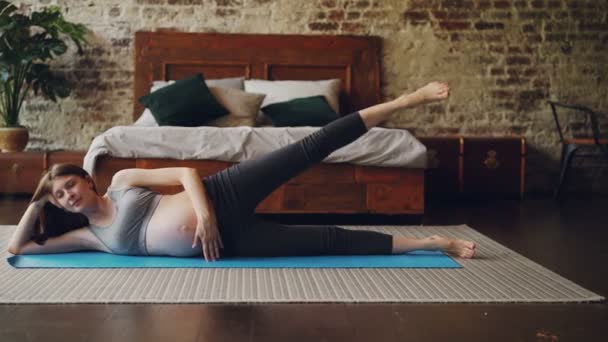 Chica bastante embarazada está haciendo ejercicios deportivos levantando la pierna acostada en el piso del dormitorio en la estera de yoga con sala de estilo loft y muebles de madera en el fondo . — Vídeo de stock