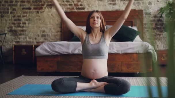 Szép terhes lány is meditál, Lótusz póz jóga mat, emeli a karját, majd üzembe pálmák, a térd és pihentető ül otthon. Jó közérzet és a terhesség koncepció. — Stock videók