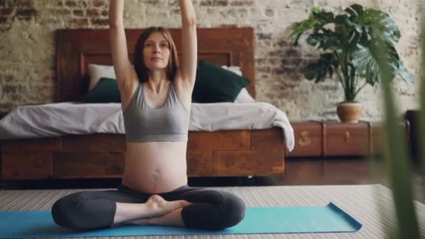 Bela mãe-a-ser está praticando hatha ioga no apartamento moderno sentado no tapete e vestindo roupas esportivas maternidade. Menina está sentada na posição de lótus e levantando os braços . — Vídeo de Stock