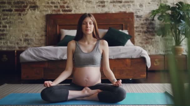 Jolie brunette enceinte mère se relaxe en position lotus sur le tapis sur le sol en pratiquant le hatha yoga avec les mains sur les genoux. Bel intérieur avec lit et plantes est visible . — Video
