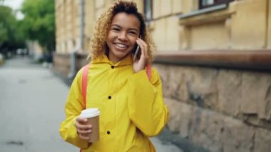 Güzel Afrikalı-Amerikalı kız cep telefonu üzerinde konuşurken ve cadde boyunca şehirde yürüyüş ve zevk konuşma gülüyor take away kahve holding.