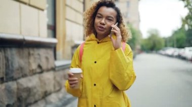Oldukça Afrikalı-Amerikalı kadın gitmek kahve içme ve cep telefonu parlak ceket ilkbaharda şehirde yürüyüş sohbet. Modern teknoloji ve millennials kavramı.