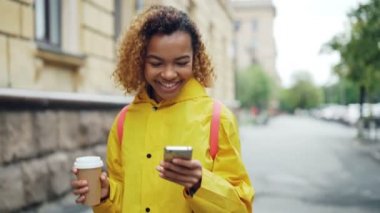 Mutlu Afrikalı-Amerikalı kadın ekran dokunaklı ve açık havada gitmek kahve ile güzel şehirde yürüyüş gülümseyen smartphone kullanıyor. Modern yaşam tarzı ve iletişim kavramı.